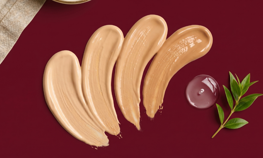 Four liquid foundation cream swatches displayed horizontally next to a transparent skincare serum droplet and a green leaf stem on a maroon backdrop