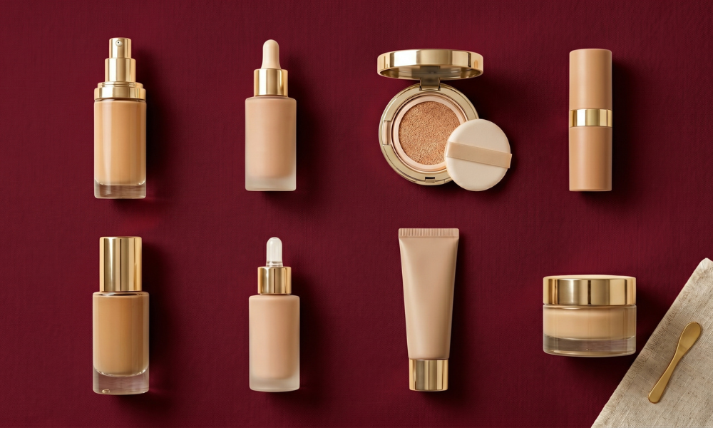 A premium cosmetics flat lay showcasing eight luxury foundation bottles cushion compacts and skincare cream jars with gold accents on a crimson background